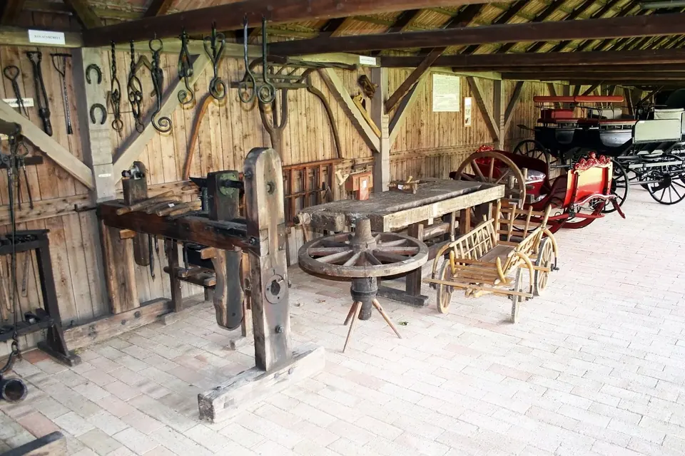 Atelier de charron reconstitué sous une charpente en bois : établis, outils suspendus, roues en cours de fabrication et voitures anciennes au fond