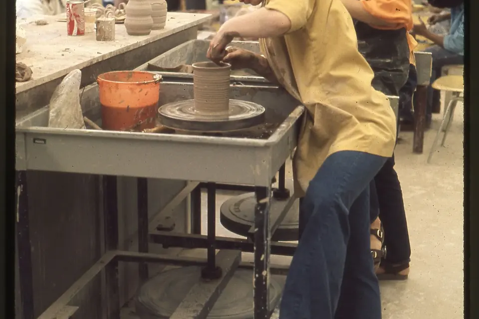 Une personne façonne un pot d'argile sur un tour de potier, dans un atelier où des pièces sèchent sur l'établi