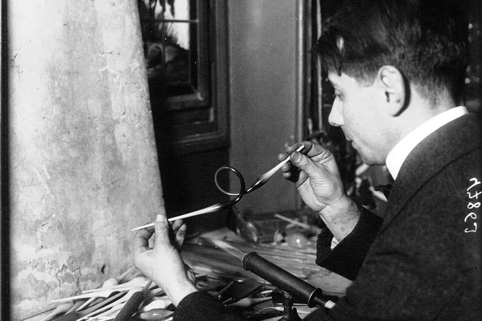 Photographie de presse ancienne en noir et blanc : un verrier assis façonne une fine pièce de verre à la flamme d'un chalumeau, pince à la main